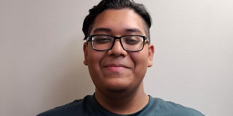 Shazadul Islam with short dark hair and glasses smiles slightly while standing in front of a plain, light-colored wall, wearing a teal t-shirt.