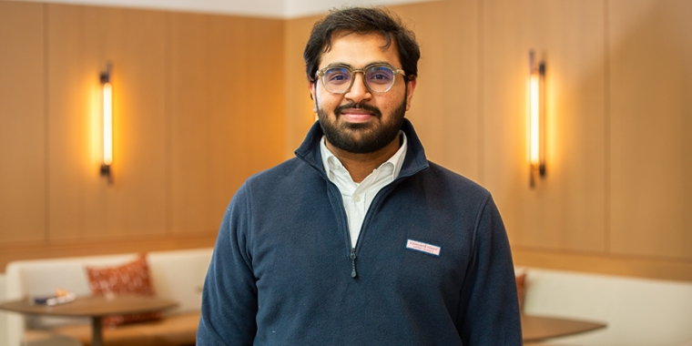 Subhan Tariq with glasses and a beard, wearing a navy pullover and white collared shirt, stands indoors in a warmly lit room with light wooden walls and cushioned seating in the background.