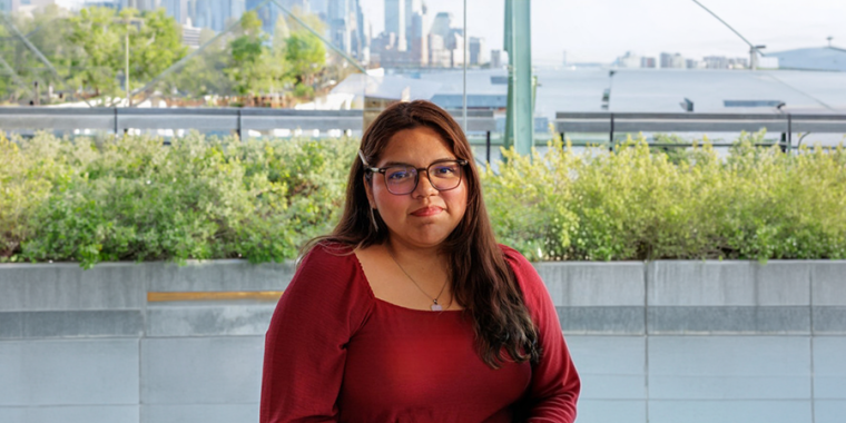 Yadhira Vivaldo Garcia with long brown hair and glasses, wearing a red blouse, sits outdoors in front of greenery and glass windows, smiling slightly at the camera.