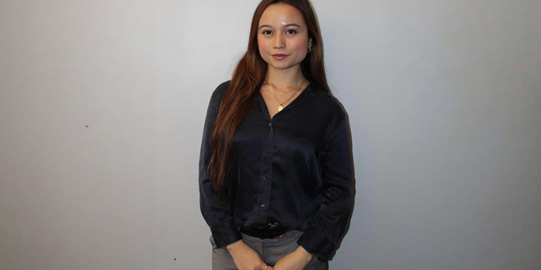 Andrea Quinonez with long brown hair, wearing a dark blouse and gray pants, stands against a plain light gray background with her hands clasped in front of her, looking at the camera.