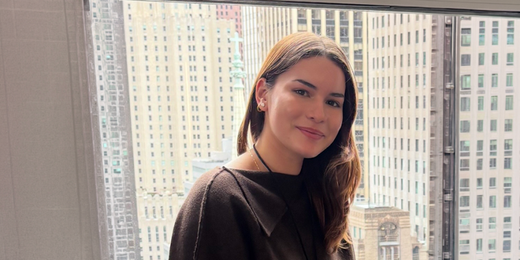 Mariana Gonzalez with long brown hair, wearing a dark top, smiles while sitting indoors in front of a large window with a New York City skyline of tall buildings in the background.