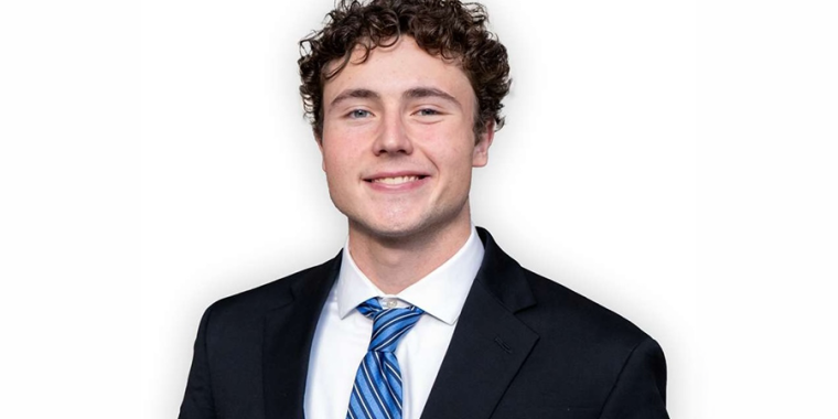 Ron Kullashi with curly brown hair, wearing a black suit, white shirt, and blue striped tie, smiles at the camera against a plain white background.
