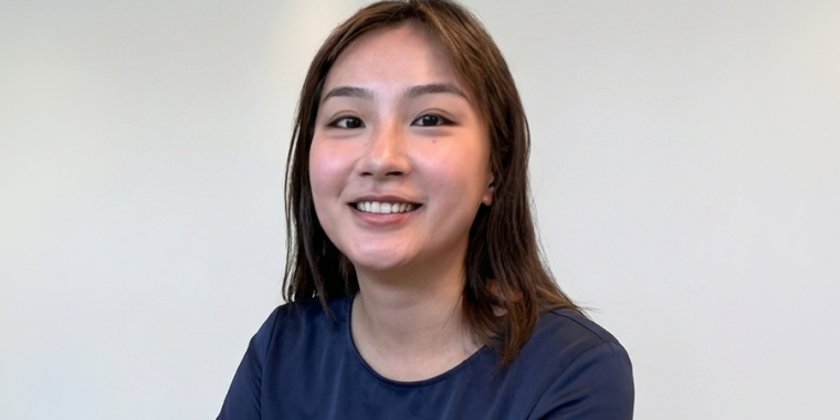 Shasuna Lin with straight brown hair and a navy blue shirt smiles at the camera against a plain, light-colored background.