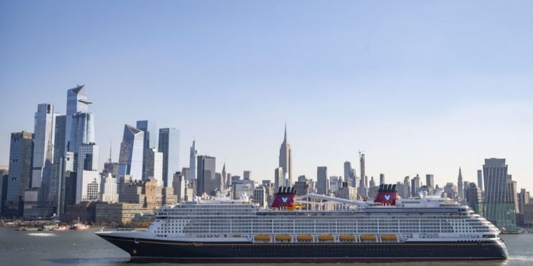Large Disney Cruise ship sails on Hudson River with New York City skyline in the background, featuring tall modern skyscrapers and clear blue sky.