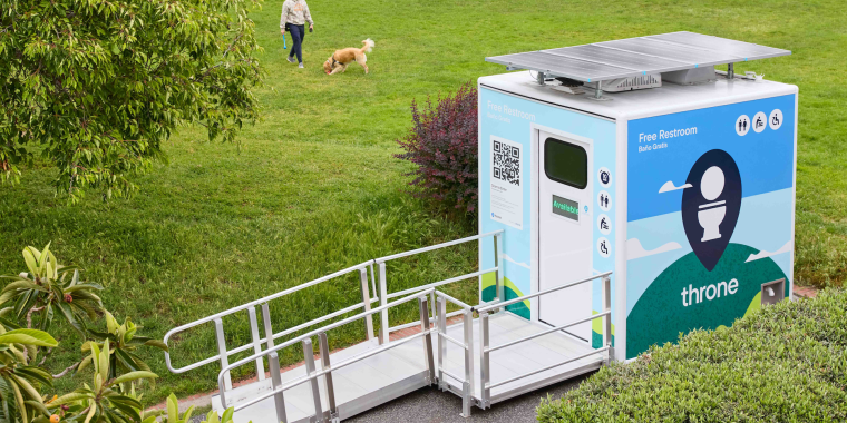 A solar-powered public restroom labeled ”Throne“ sits in an NYC park with a ramp; a person walks a dog in the grassy area nearby. Photo credit: Megan Bayley.