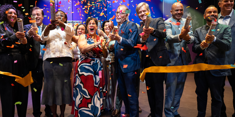 A group of people celebrates at a ribbon-cutting ceremony, releasing colorful confetti. They are smiling, clapping, and dressed in formal attire on a decorated stage.