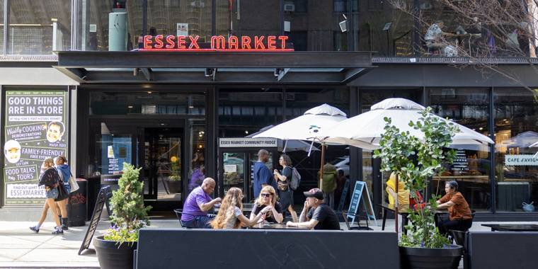 People sit at outdoor tables under umbrellas in front of Essex Market. Some are eating and chatting. Large potted plants line the seating area, and the markets glass entrance and signage are visible behind them.