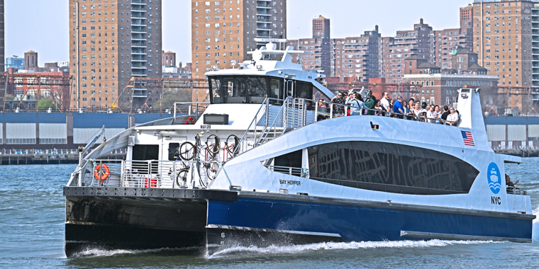 A blue and white NYC Ferry boat, the Bay Hopper, travels across the water with passengers on the upper deck and bicycles racked on the front.