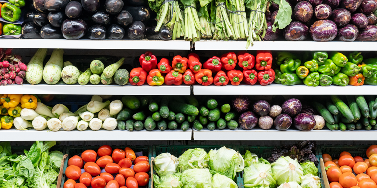 Shelves of fresh vegetables in a grocery store, including eggplants, leafy greens, peppers, cucumbers, tomatoes, and cabbages, all neatly arranged in rows.