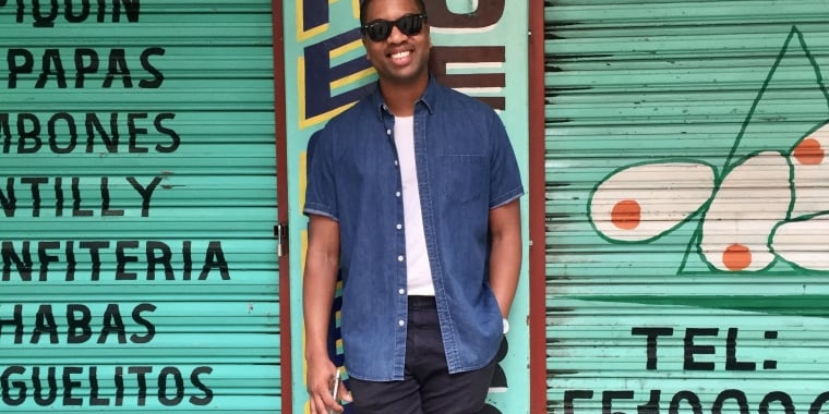 Erich Bilal wearing sunglasses, a blue shirt, and black pants stands smiling in front of a teal shutter with Spanish words painted on it. He holds a drink bottle in one hand. The shutter features abstract shapes and a phone number.