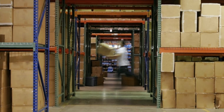 A person in motion, carrying boxes in a warehouse aisle filled with stacked brown cardboard boxes on metal shelves. The floor is concrete, and the shelves extend high, suggesting a large storage facility.