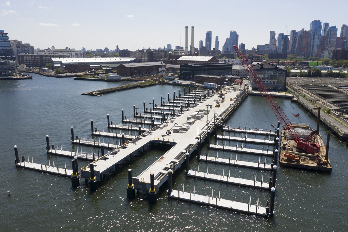 Aerial view of the Brooklyn Navy Yard Ferry pier construction with an L-shaped dock structure extending into the water. Several empty dock slips are visible. A red crane and construction materials are on-site. Cityscape and industrial buildings are in the background.