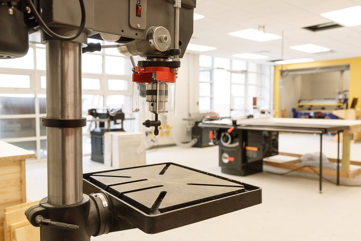 An industrial workshop with a large drill press in the foreground. The room is spacious and well-lit with large windows. In the background, there is a workbench and various tools and equipment scattered around. The walls are yellow and white. Photo by Kreg Holt/NYCEDC. An industrial workshop with a large drill press in the foreground. The room is spacious and well-lit with large windows. In the background, there is a workbench and various tools and equipment scattered around. The walls are yellow and white. Photo by Kreg Holt/NYCEDC.