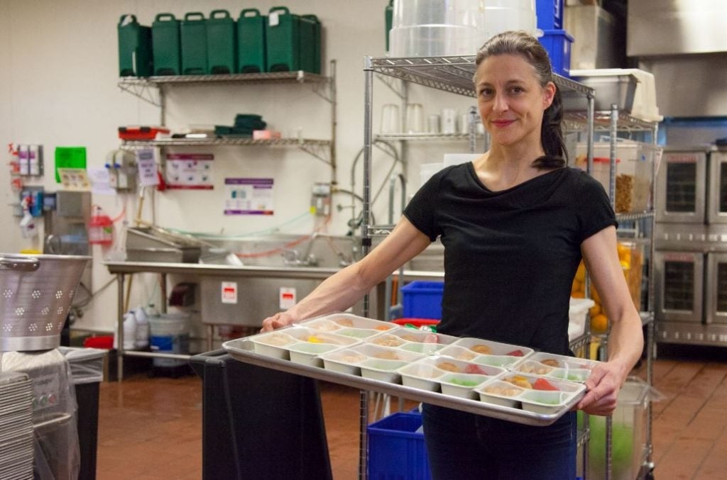 A person in kitchen of Little Green Gourmets at La Marqueta Market holds a tray of colorful, portioned meals. Shelves filled with kitchen supplies and ingredients are visible in the background. The setting is clean and organized. A person in kitchen of Little Green Gourmets at La Marqueta Market holds a tray of colorful, portioned meals. Shelves filled with kitchen supplies and ingredients are visible in the background. The setting is clean and organized.