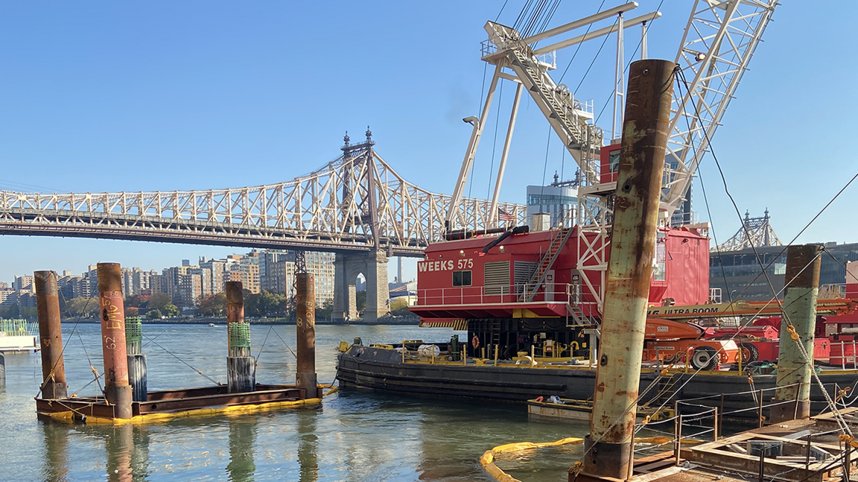Construction site of Offshore Wind NYC Waterfront Pathways Program, showing a red crane barge on a river with the Queensboro Bridge in the background. Several pilings are in the water, and buildings line the opposite riverbank under a clear blue sky. Construction site of Offshore Wind NYC Waterfront Pathways Program, showing a red crane barge on a river with the Queensboro Bridge in the background. Several pilings are in the water, and buildings line the opposite riverbank under a clear blue sky.