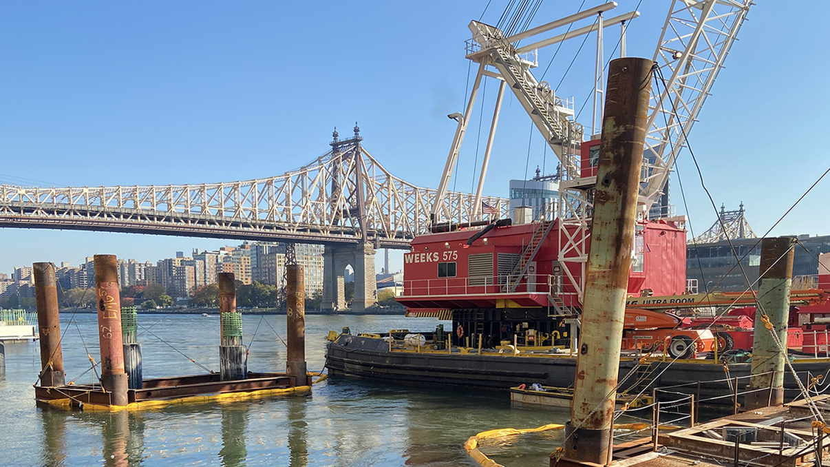 Construction site of Offshore Wind NYC Waterfront Pathways Program, showing a red crane barge on a river with the Queensboro Bridge in the background. Several pilings are in the water, and buildings line the opposite riverbank under a clear blue sky.