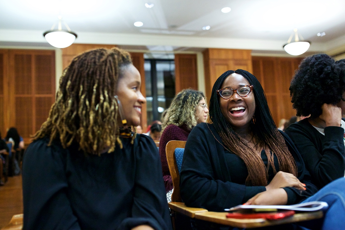 Two female participants at a Ask for More salary negotiation seminar. They are sitting on chairs in a room, smiling and talking. One has long braided hair and glasses, while the other has curly hair. They are surrounded by other people seated in a lecture hall with wooden paneling and ceiling lights.