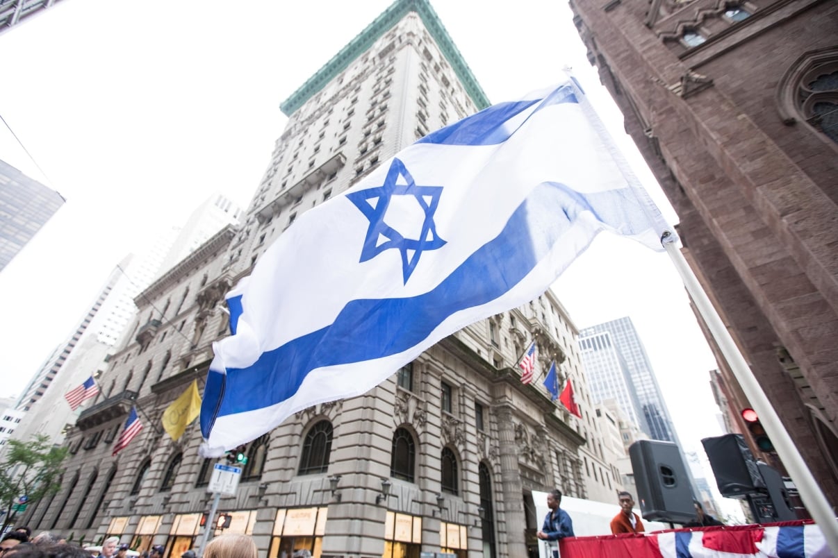 Israeli flag waving in a city street filled with tall buildings and a few people. Other flags, including an American one, are visible. The scene includes loudspeakers and parts of old architecture. Israeli flag waving in a city street filled with tall buildings and a few people. Other flags, including an American one, are visible. The scene includes loudspeakers and parts of old architecture.