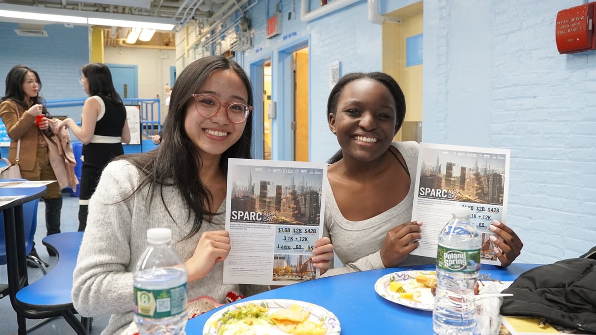 Two people smiling and holding flyers while seated at a table with food. The flyers display text and city images. Other individuals are visible in the background, and the setting appears to be a bright, casual indoor space.