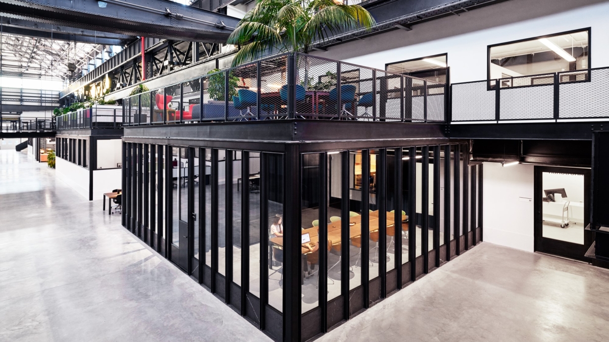 Interior image of New Lab showing a spacious modern office with high ceilings and an industrial design. Features glass-walled conference room, upstairs seating area with plants, and open spaces with natural light. Photo courtesy of New Lab.