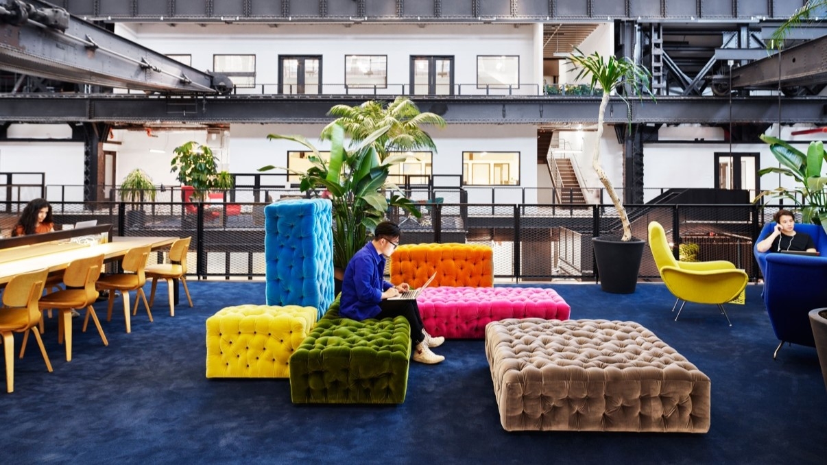 Interior shot of New Lab showing a modern office space, with vibrant and tufted seating in various colors on a blue carpet. Wooden table and chairs are on the left, while plants add greenery. A man in a blue jacket is working on a laptop. Open industrial design with offices in the background.