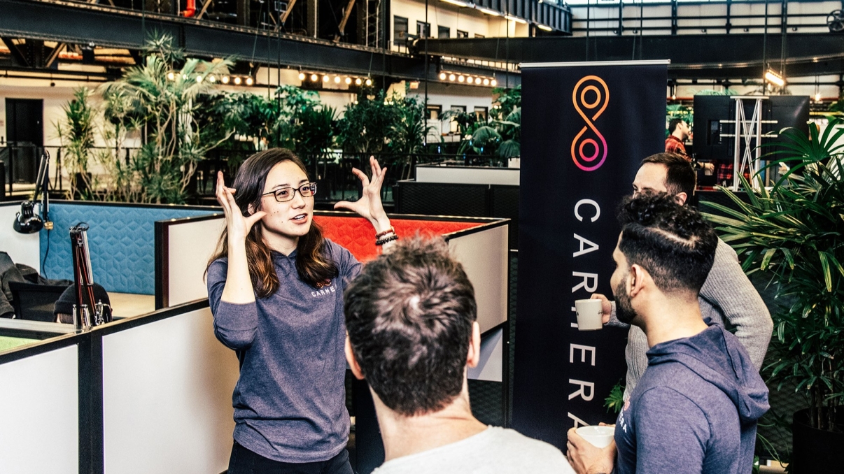 A woman energetically talks with three men in modern office space at New Lab. They are casually dressed, and two men hold coffee cups. Lush greenery decorates the space, and a banner with a colorful infinity logo stands nearby. Photo courtesy of New Lab.