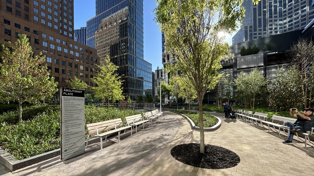 Abolitionist Place Park in Brooklyn with green trees, people sitting on benches, and high-rise buildings in the background.