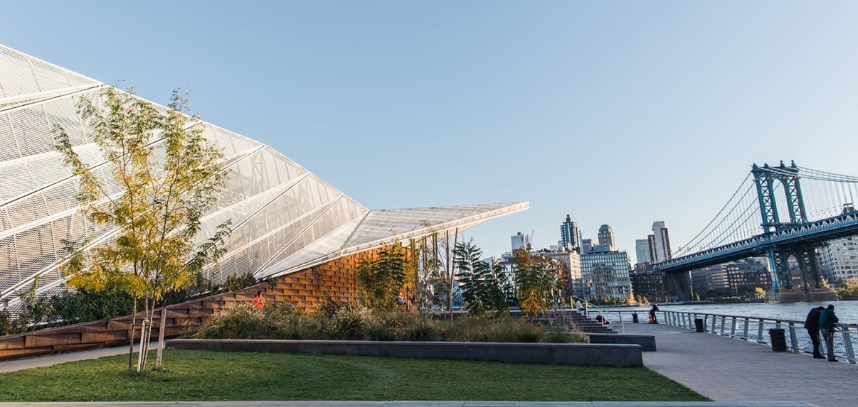 Modern building with angled roof and lush landscaping on Pier 35 on the East River, overlooking a city skyline and a large bridge.