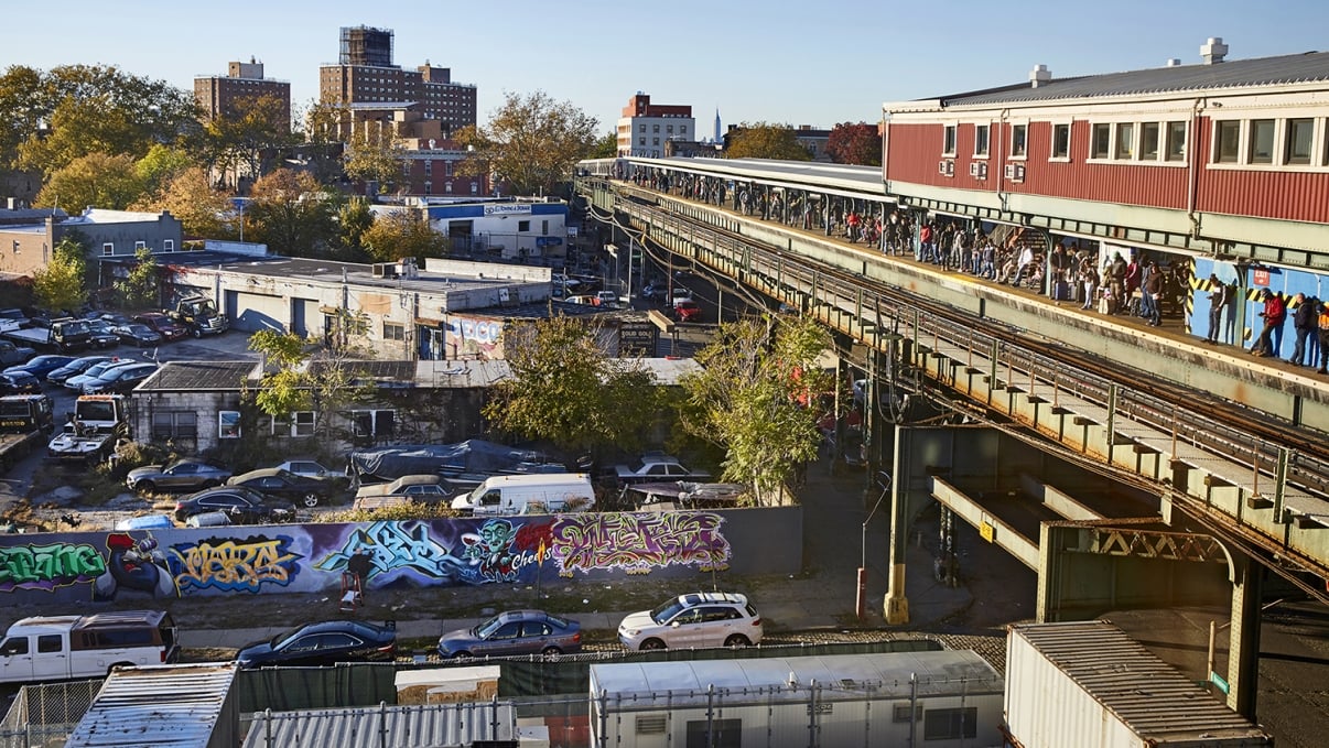 An elevated train station bustling with people above a graffiti-covered wall and parked cars. Buildings and trees are in the background.