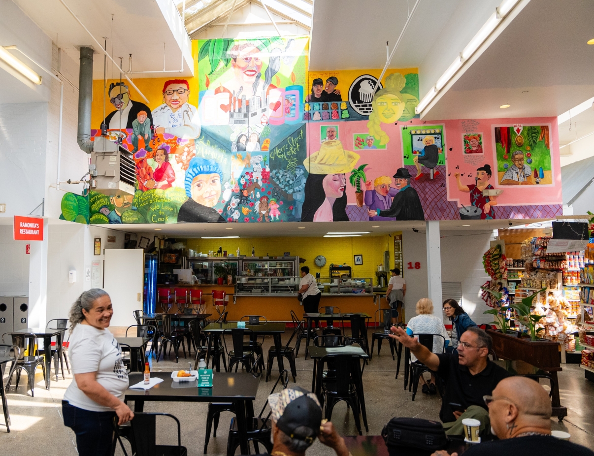 People sit and talk at tables inside a bright, casual market or café. A colorful mural with portraits and vibrant scenes covers the upper wall above the counter, adding artistic flair to the lively indoor space.