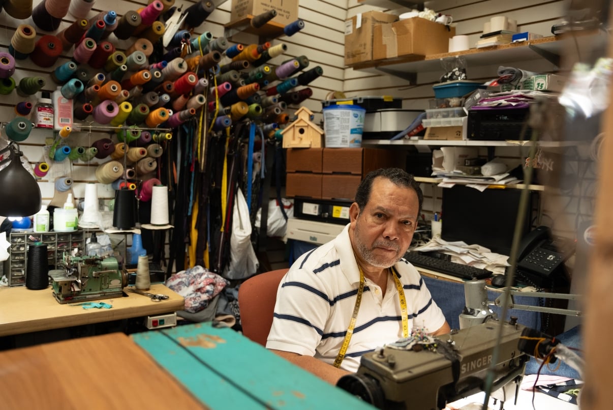A man with short hair and a beard sits at a sewing machine in a workshop, smiling. He wears a striped shirt and a yellow tape measure around his neck. Shelves with various supplies and tools are in the background.
