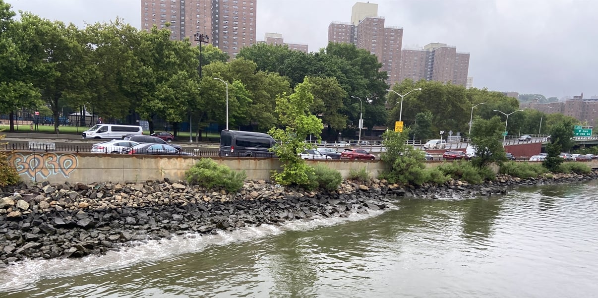 North Harlem waterfront with a rocky shoreline, a road with traffic, trees, and high-rise buildings in the background under a cloudy sky.