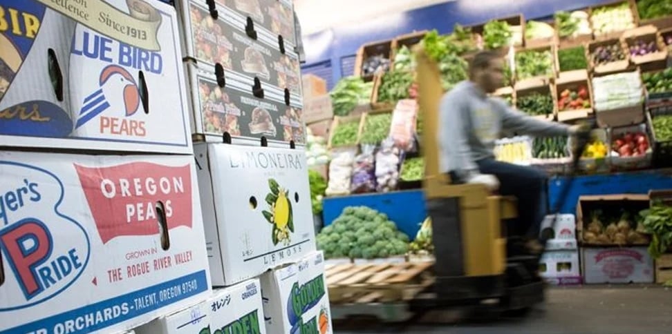 A worker on a forklift moves past stacked fruit boxes in Hunts Point Produce Center, with a variety of vegetables displayed on shelves in the background.