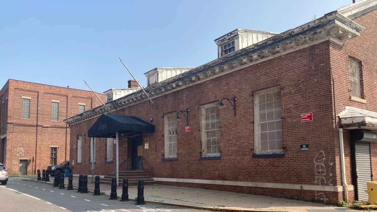 Brick Bush Terminal Café Building with a black awning, barred windows, and a small loading dock. Street in foreground with bollards and a bike lane. Blue sky above.