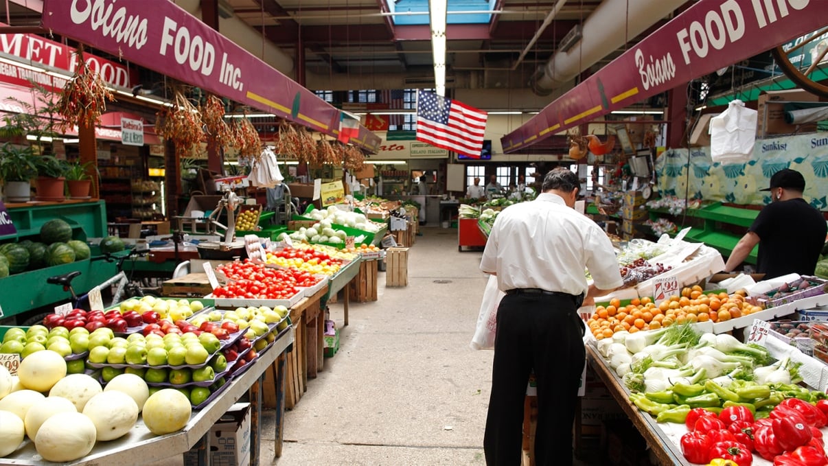 A bustling indoor market with a variety of fresh produce on display. Stalls are filled with colorful fruits and vegetables, and shoppers are browsing. An American flag hangs above, and the market has vibrant, lively signage.