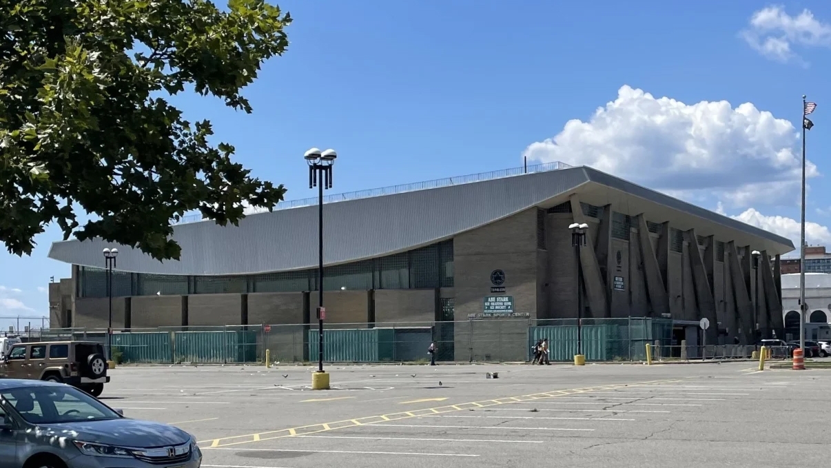 A large arena building, Abe Stark Skating Rink, with a unique architectural design featuring angular walls and a flat roof. It&#039;s surrounded by an empty parking lot in Coney Island under a clear blue sky with some fluffy clouds. A tree branch is visible in the foreground.