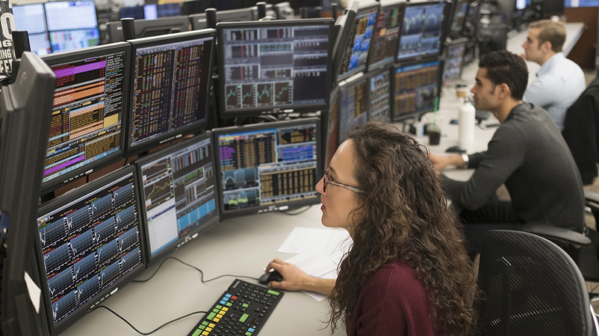 People working at desks filled with multiple computer monitors displaying financial data and charts. A woman with curly hair is focusing on her screens, while two men are working in the background. The scene is busy and tech-oriented.