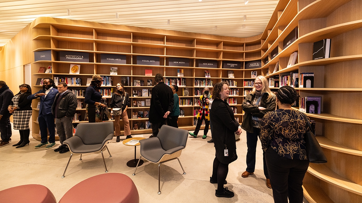 People are gathered in a modern library space with curved wooden shelves filled with books. Several small groups are conversing while others browse the selection. The area features comfortable seating and warm lighting.