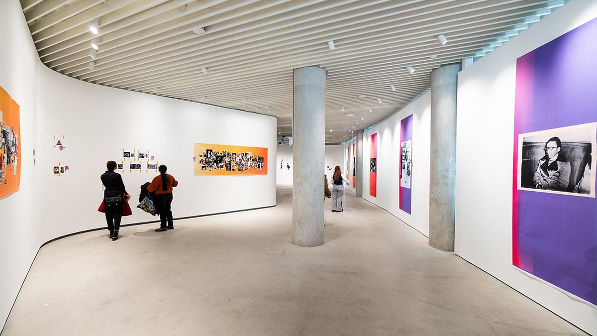 A modern art gallery with white curved walls featuring colorful displays and photo collages. Several people are observing the artwork, with a focus on a large photo of a seated person on the right wall. The ceiling has a striped pattern.