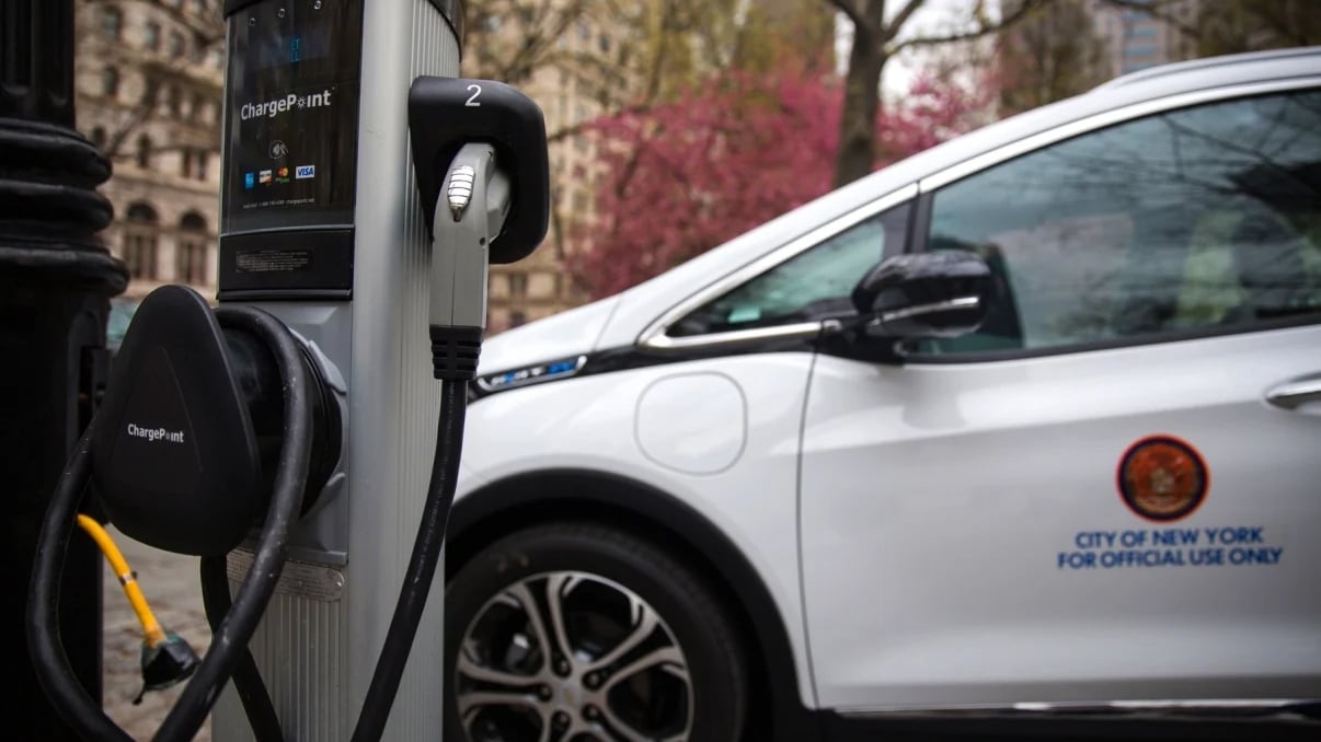 An electric vehicle charging at a public charging station. The vehicle is white with a City of New York for Official Use Only logo. Trees and buildings are visible in the background.