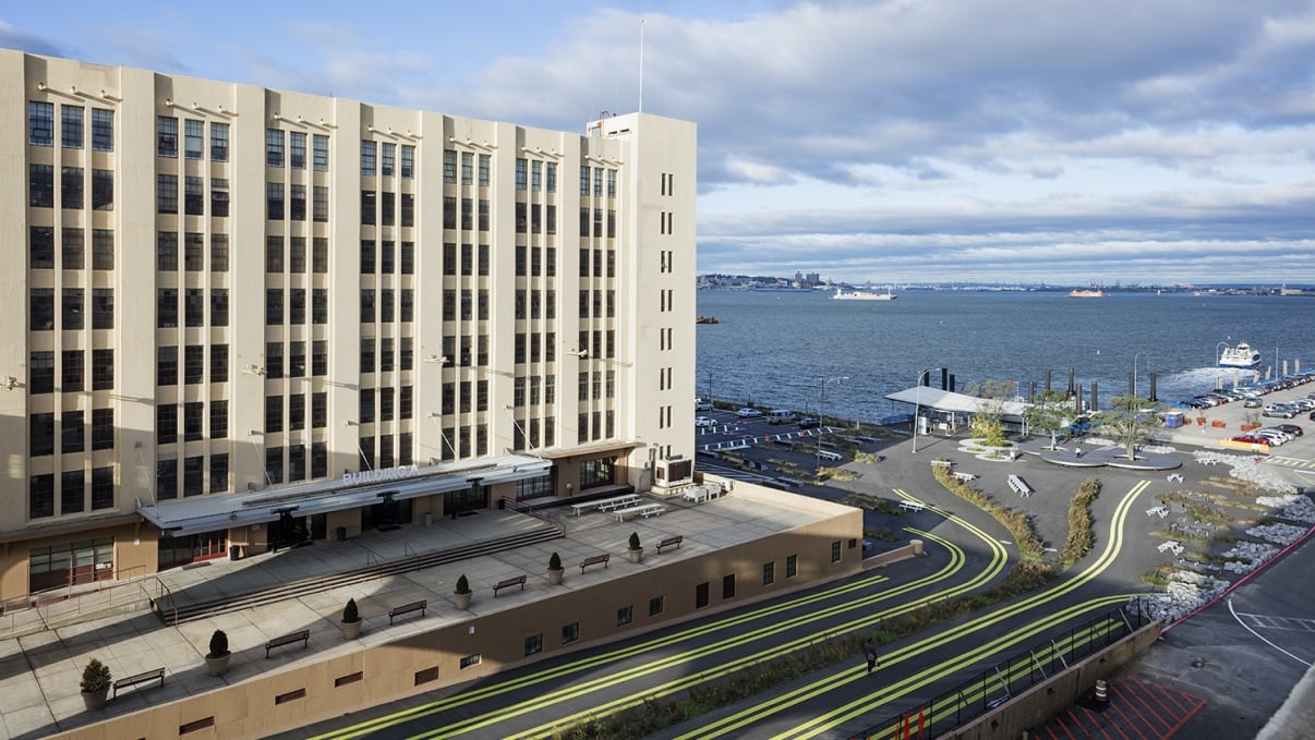 A large beige building overlooks a harbor with several boats. In the foreground, there are yellow-striped lanes and sparse vegetation. The sky is cloudy, and distant land is visible across the water.