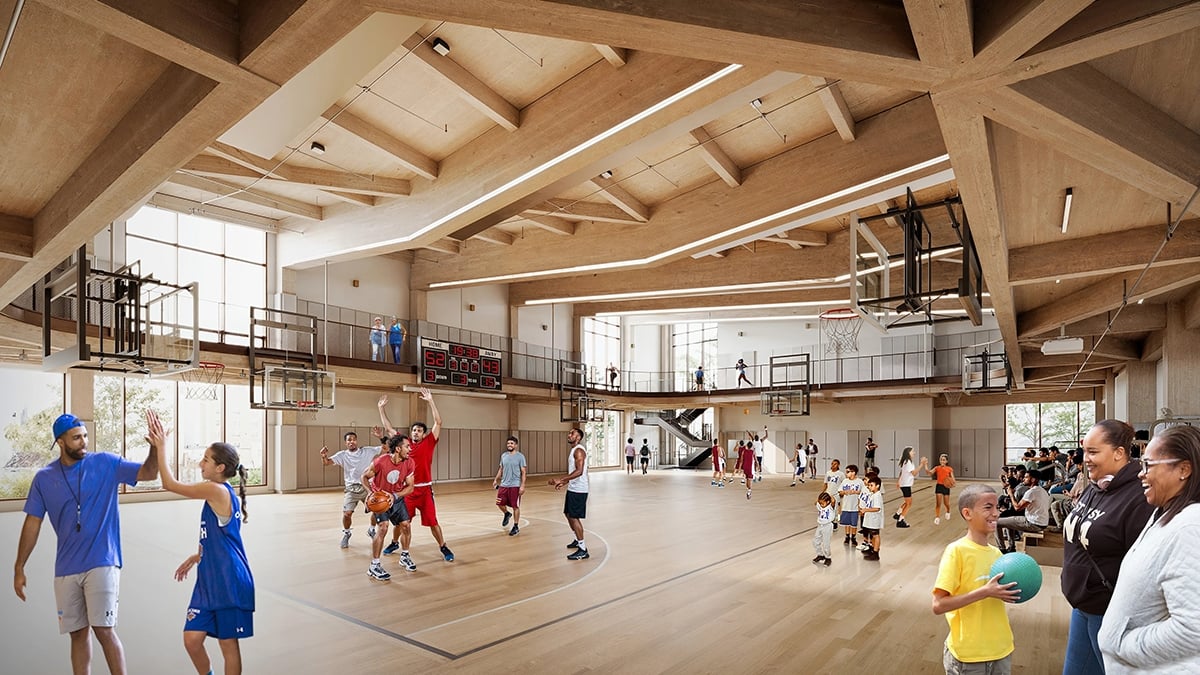 A large indoor basketball court with people playing various sports. Children and adults are engaging in activities like basketball and passing balls. Spectators are watching from a mezzanine level. The hall features wooden ceilings and large windows.