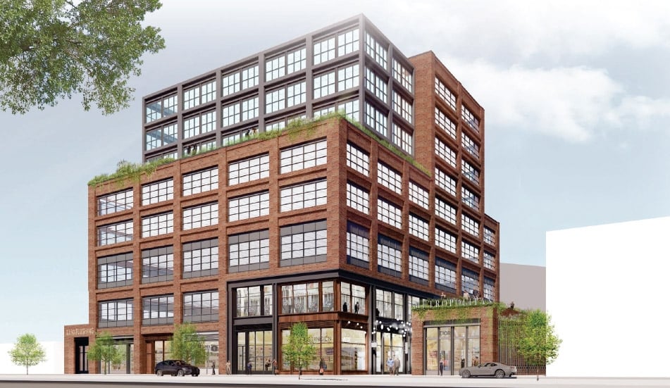 A multi-story, red-brick building with large, grid-style windows. Upper floors feature black-framed windows. Greenery lines the edges of a rooftop terrace. The ground floor has storefronts with glass doors and windows. A tree is partially visible.