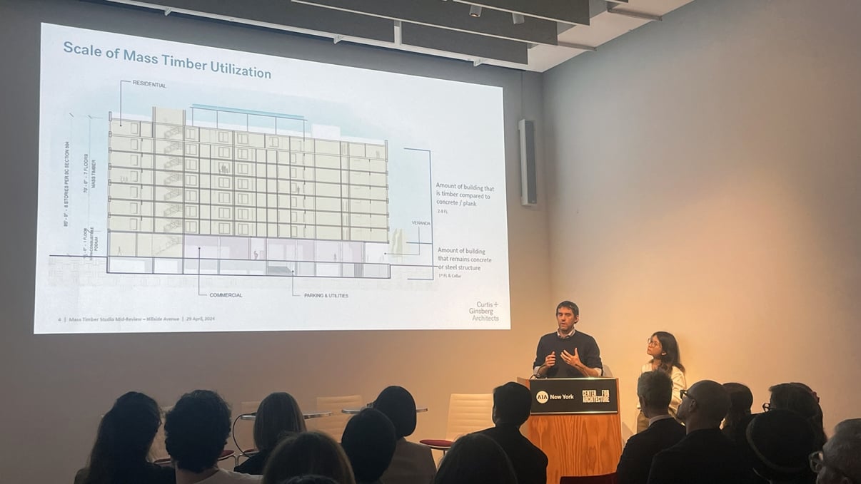 Two speakers present a slideshow on Scale of Mass Timber Utilization to an audience in a dimly lit room. The presentation includes architectural diagrams and information about timber usage.