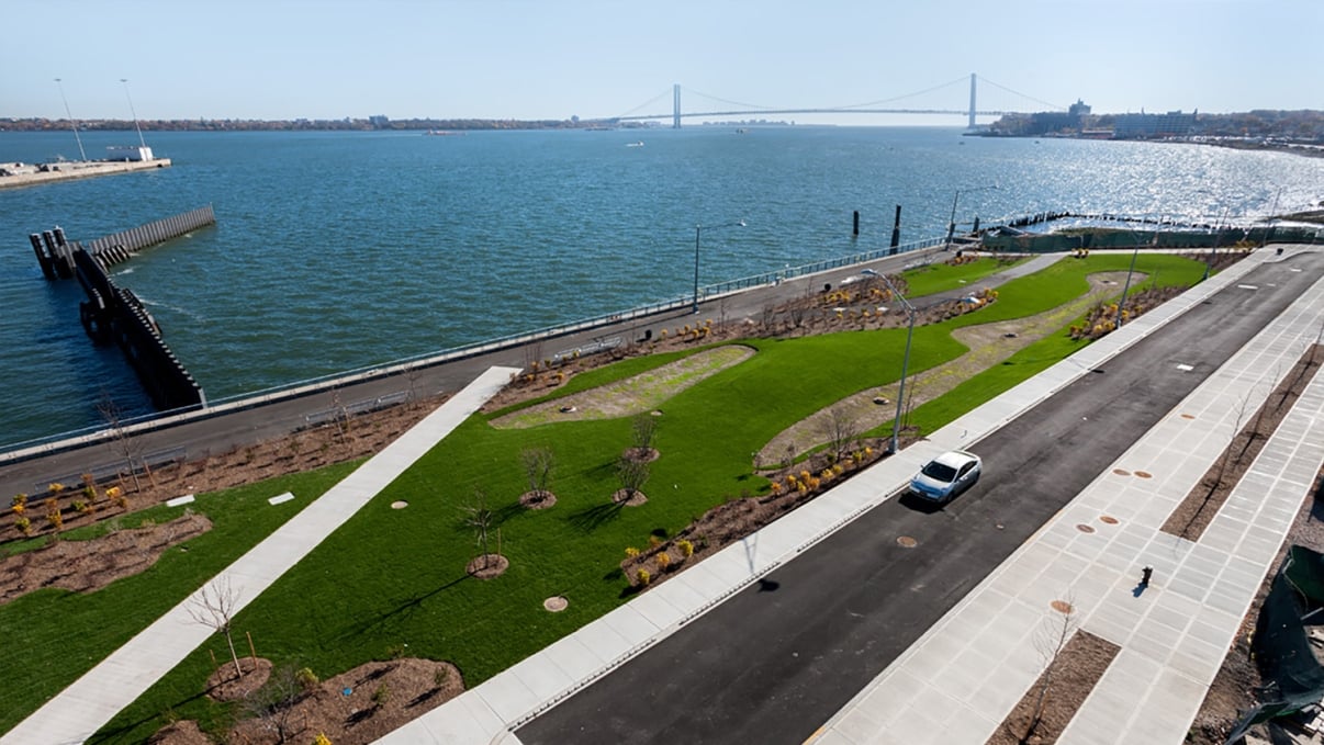 Aerial view of a waterfront park with green lawns and pathways along the shoreline. A road runs beside the park, and a bridge is visible in the background across the water. Sunny, clear skies.