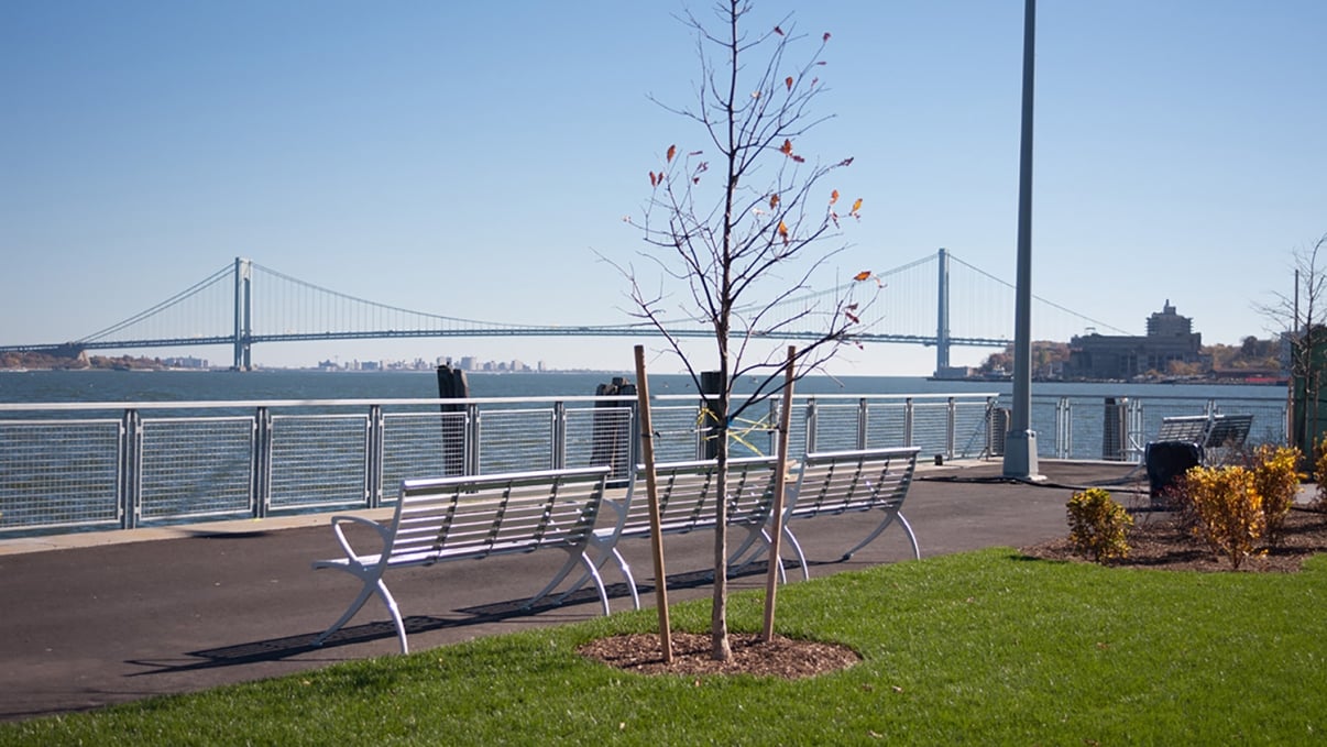 A riverside park scene with white benches and a small tree in the foreground. In the background, a suspension bridge spans across the water under a clear blue sky. A path runs alongside the river with neatly trimmed grass and shrubs.