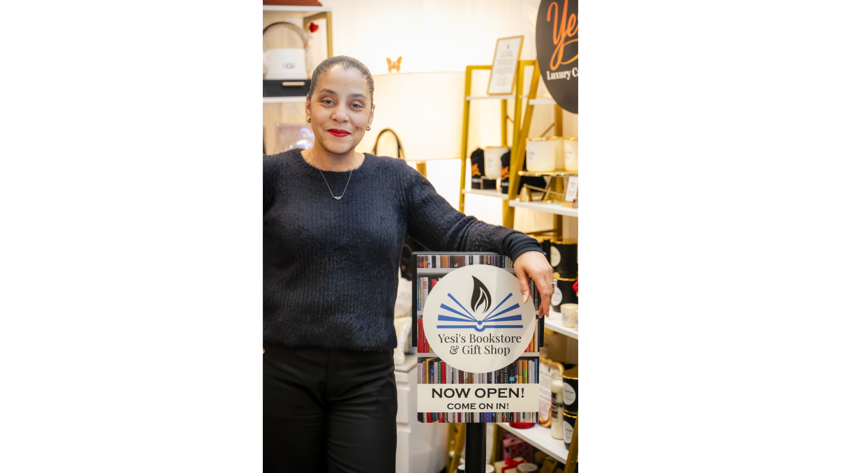 A woman smiling and standing in a bookstore, holding a sign that reads Yesis Bookstore &amp;amp; Gift Shop, NOW OPEN! COME ON IN! The store has shelves with books and gift items in the background.