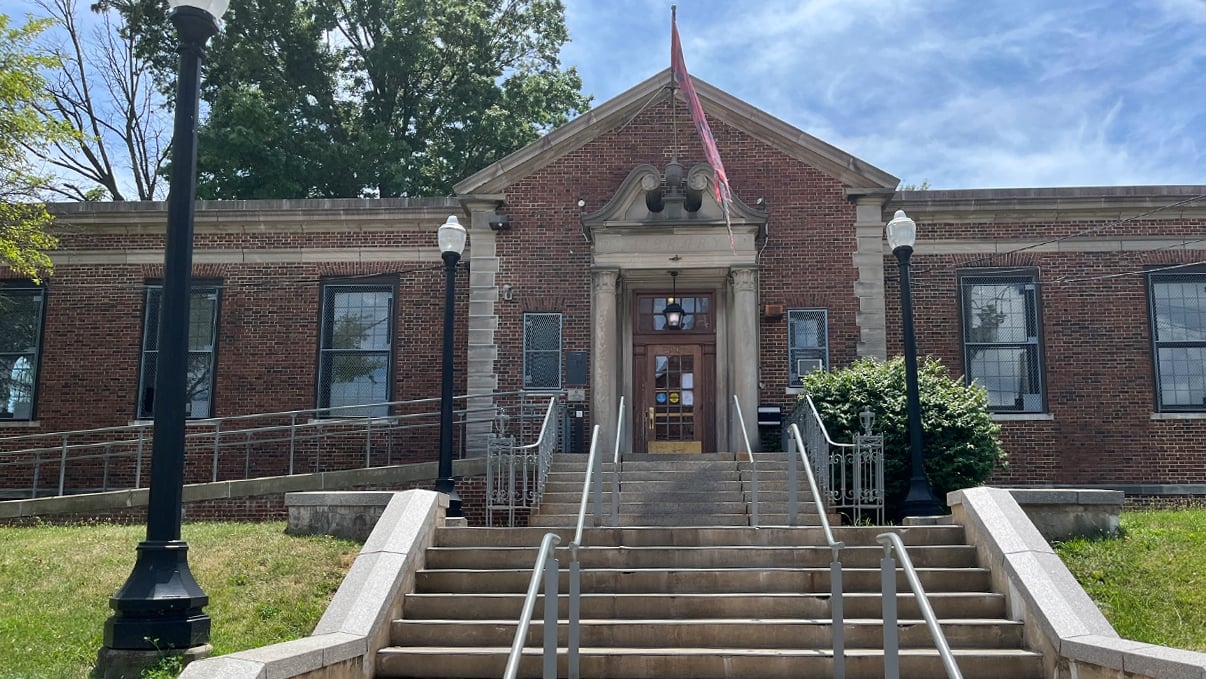 A brick building with a central entrance featuring a small porch and steps, flanked by metal railings. An American flag flies above the entrance. There are trees in the background and patches of snow on the ground.