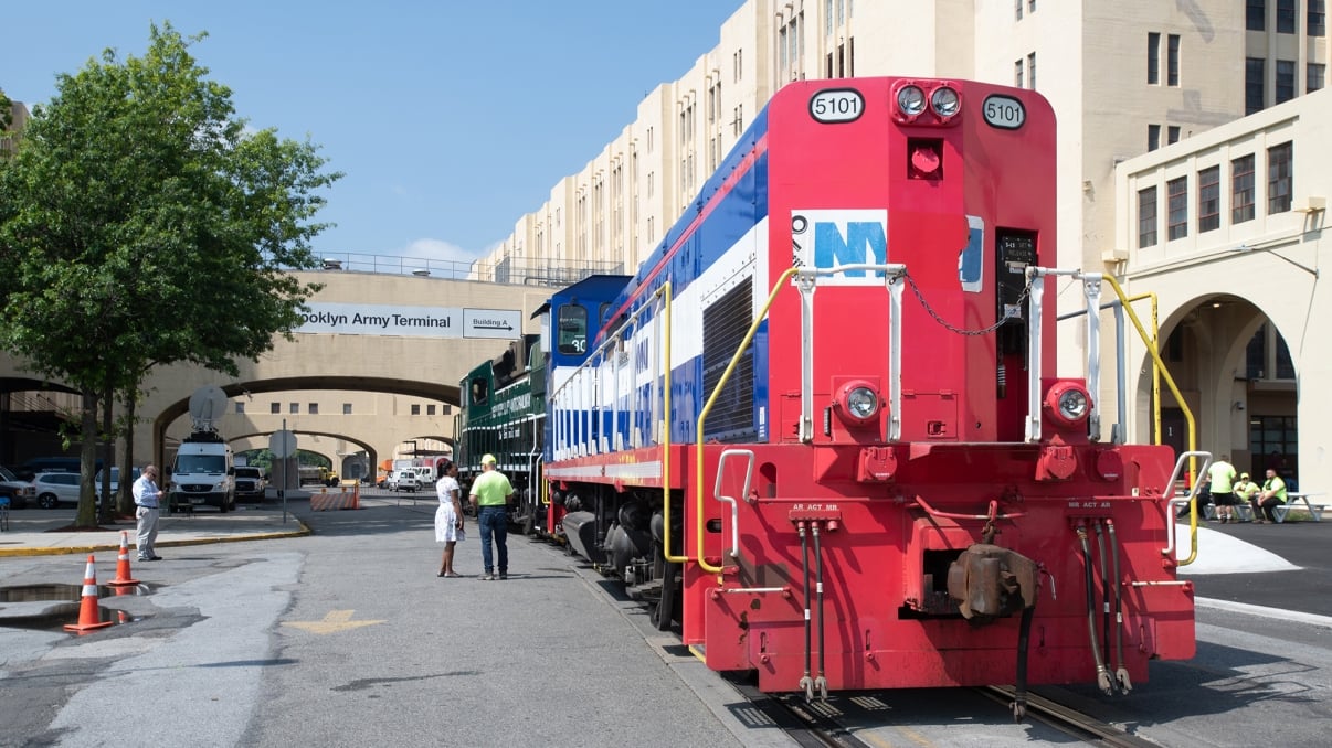 A colorful train with red, white, and blue locomotives is parked at the Brooklyn Army Terminal. Several people in safety vests stand nearby. A large building and a tree are in the background under a clear blue sky.