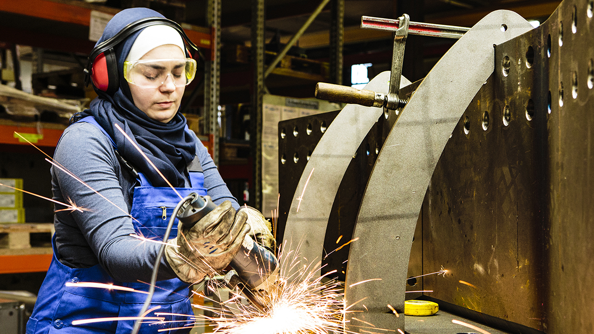 A person in a hijab and protective gear operates a grinder in a workshop, surrounded by metal and sparks. They wear gloves, safety goggles, and ear protection, focusing intently on their work.